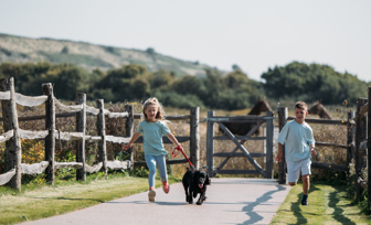 Two young children running along a path in the countryside with a black dog surrounded by fencing and a gate out to a nature field