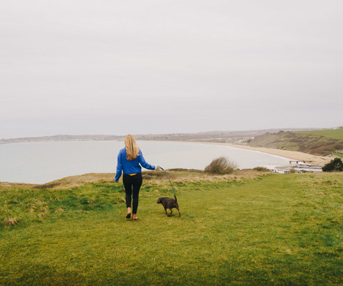 A lady walking on the south west coast path with a dog and views of Weymouth Bay