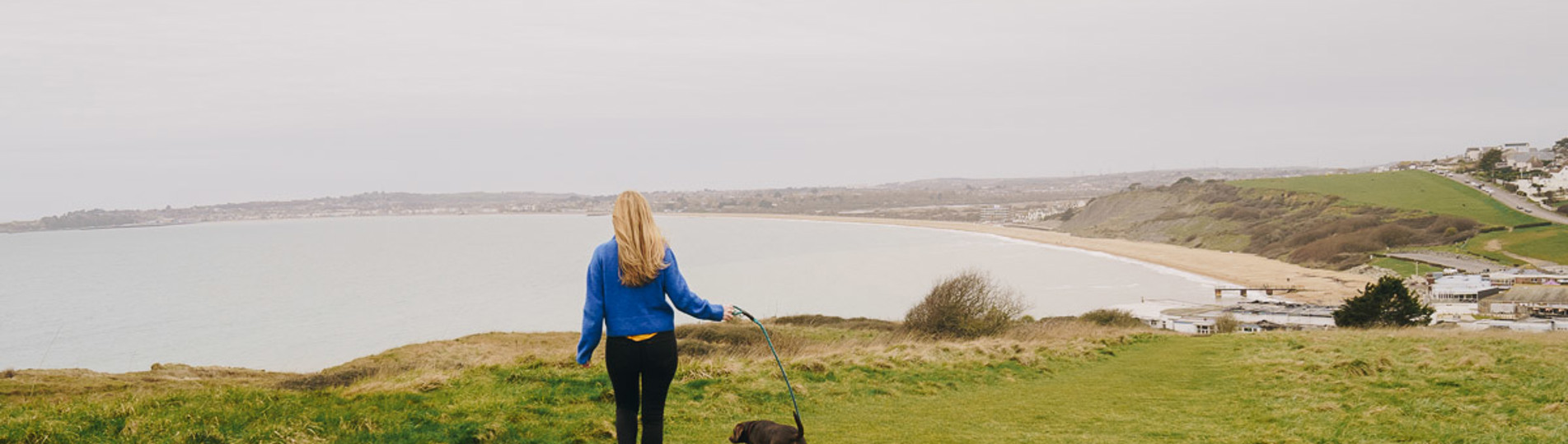 A lady walking on the south west coast path with a dog and views of Weymouth Bay