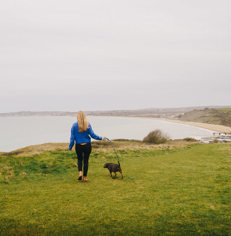 A lady walking on the south west coast path with a dog and views of Weymouth Bay
