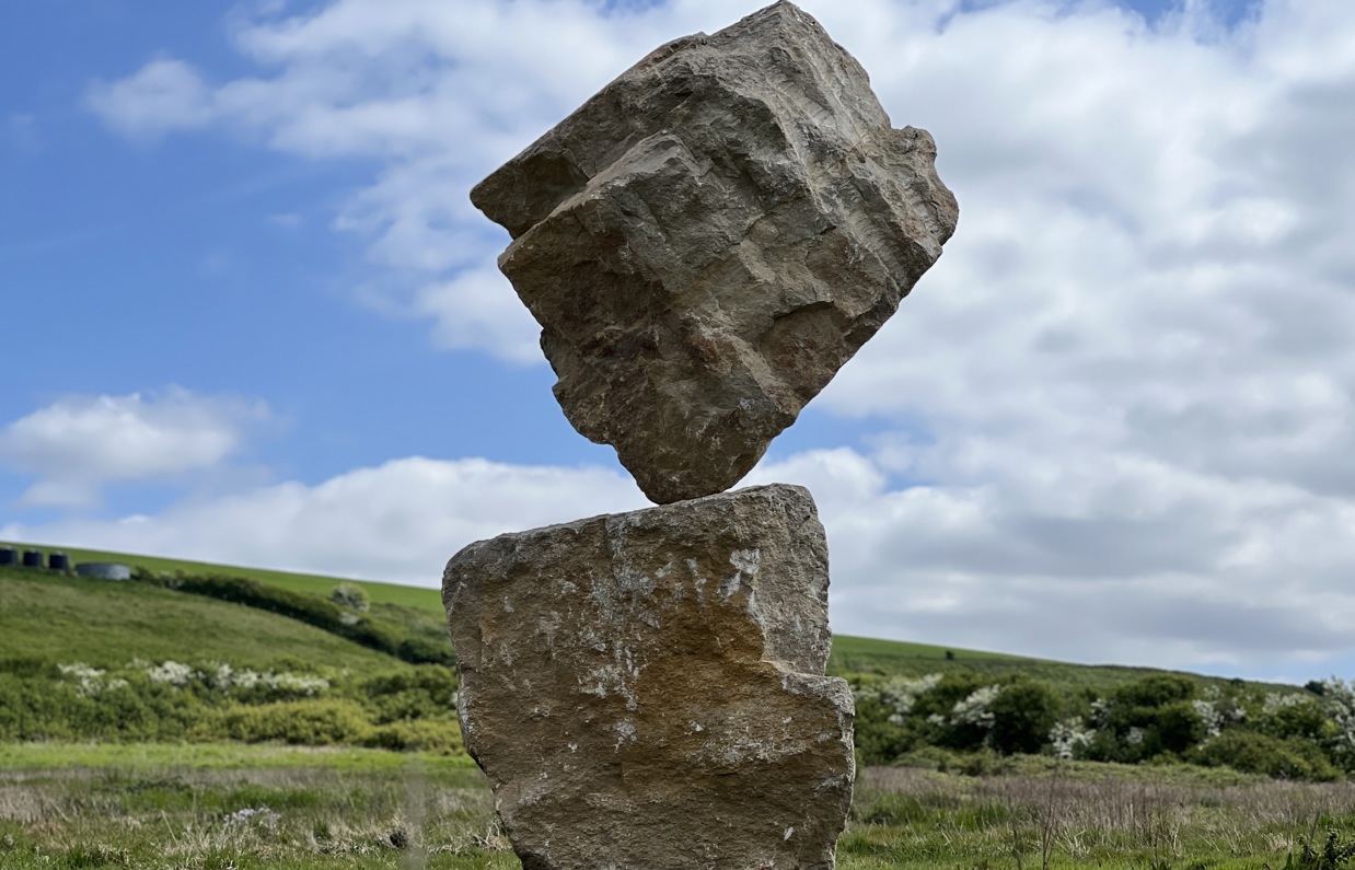 A carefully balanced stone sculpture in a countryside field with blue skies and clouds in the background