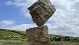 A carefully balanced stone sculpture in a countryside field with blue skies and clouds in the background