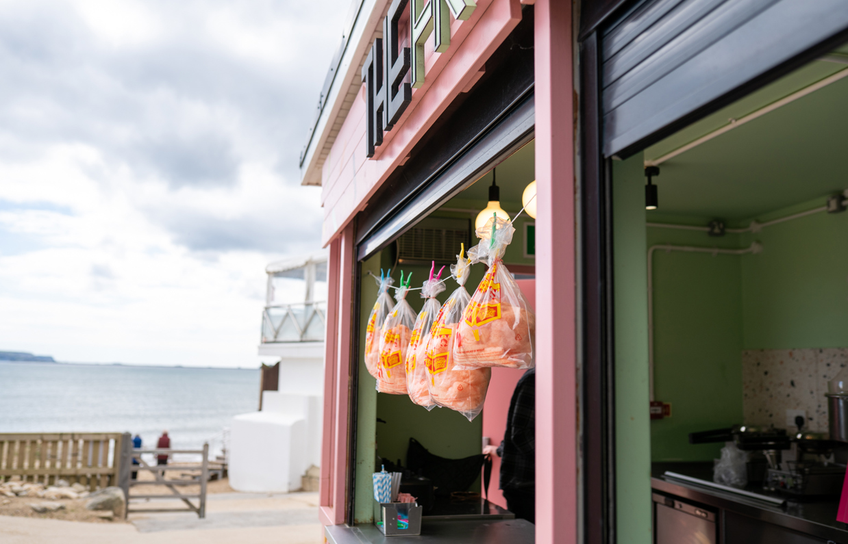 The outside of a dessert bar called The Frostbite with candyfloss hanging from the hatch and the sea in the background