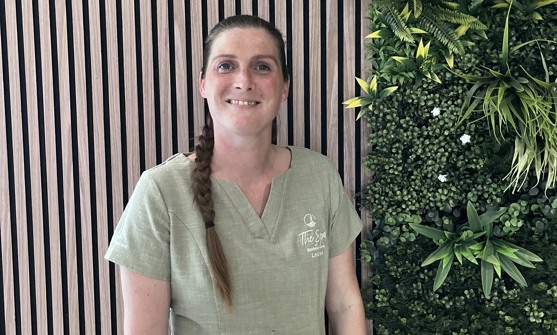 A lady smiling in spa uniform against a greenery / walnut decór background