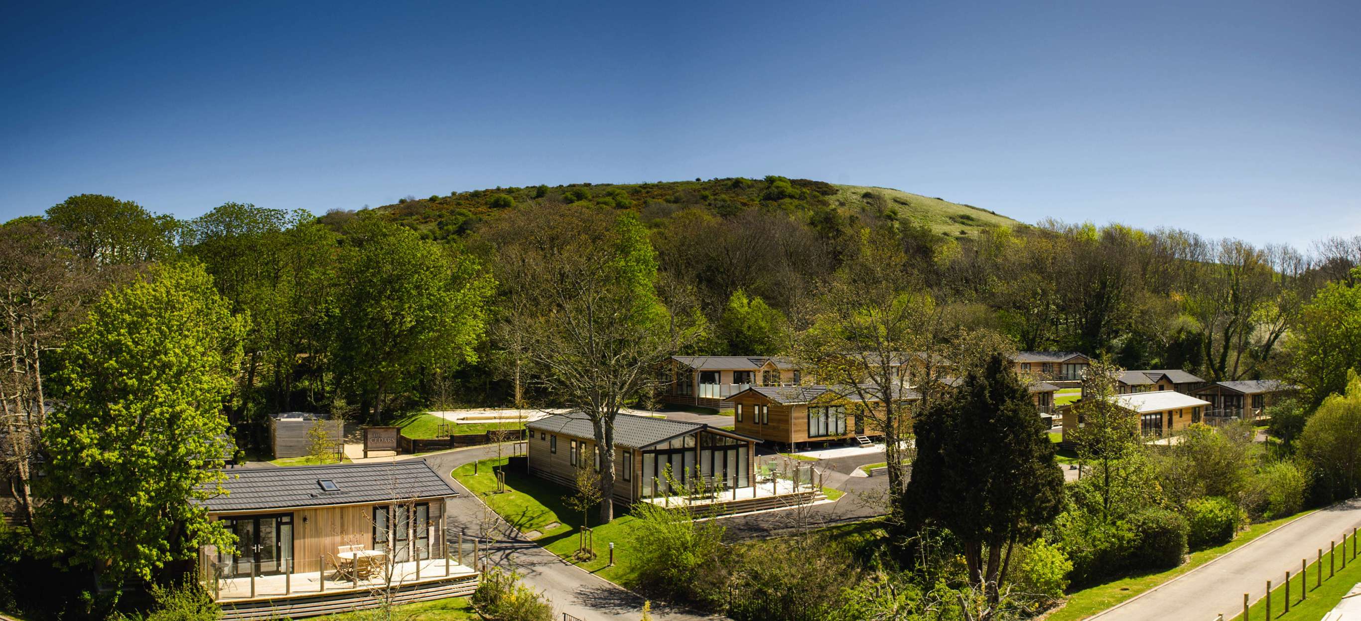Holiday lodges among woodland at the bottom of rolling countryside hills on a sunny, blue sky day