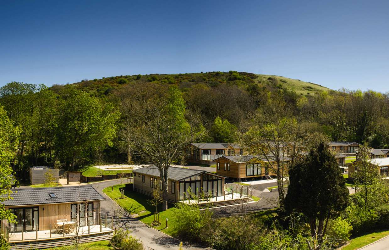 Holiday lodges among woodland at the bottom of rolling countryside hills on a sunny, blue sky day