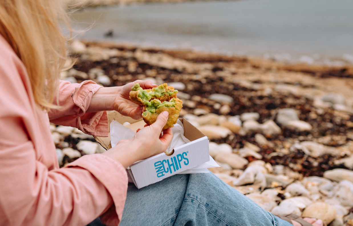 A woman sat on the beach with a fish and chip box open on her lap holding a broken open pea fritter