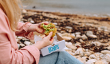 A woman sat on the beach with a fish and chip box open on her lap holding a broken open pea fritter