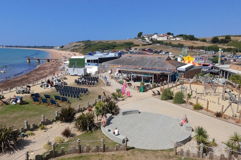 An aerial view shot of Southside Green area with deckchairs, a large TV screen, outdoor play area, food and drink outlets and Southside Funfair, all with seaviews