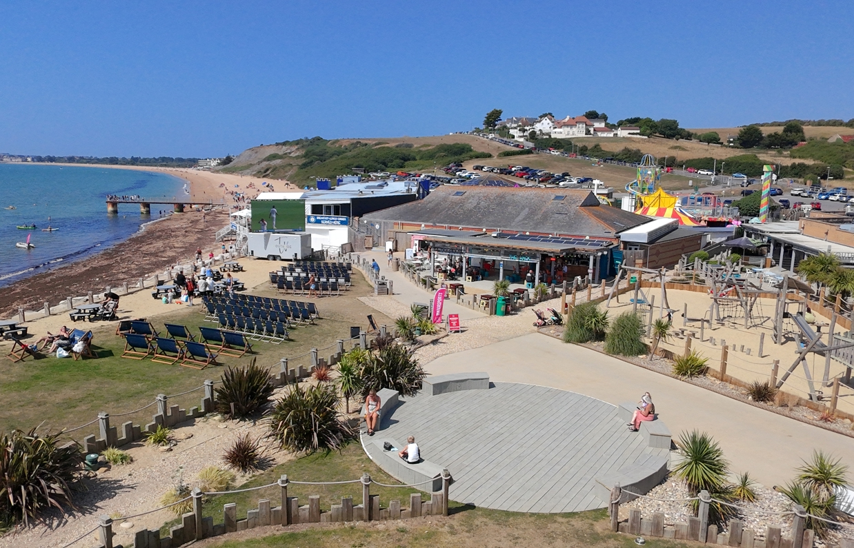 An aerial view shot of Southside Green area with deckchairs, a large TV screen, outdoor play area, food and drink outlets and Southside Funfair, all with seaviews